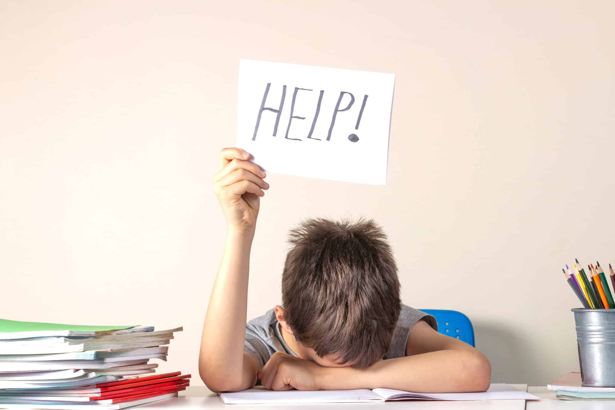 Frustrated student, head down on his desk, holds up a sign that reads Help!