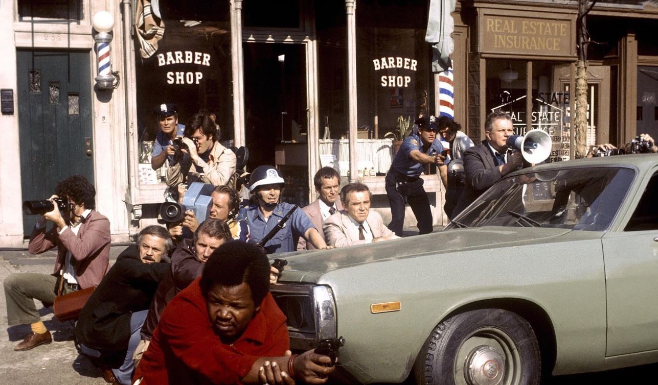A group of cops and hostage negotiators on the street huddling behind a car