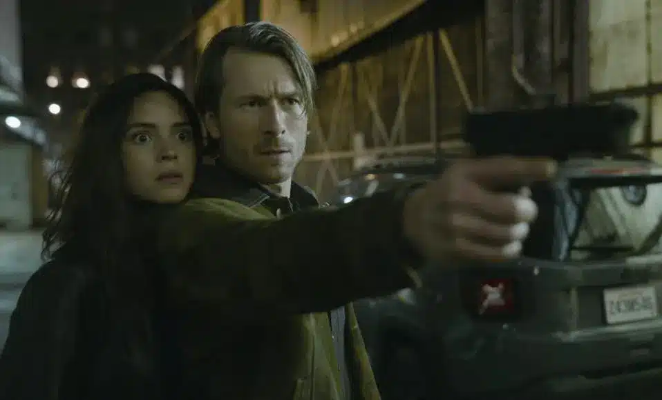 Glen Powell pointing a gun with Adria Arjona clinging to him