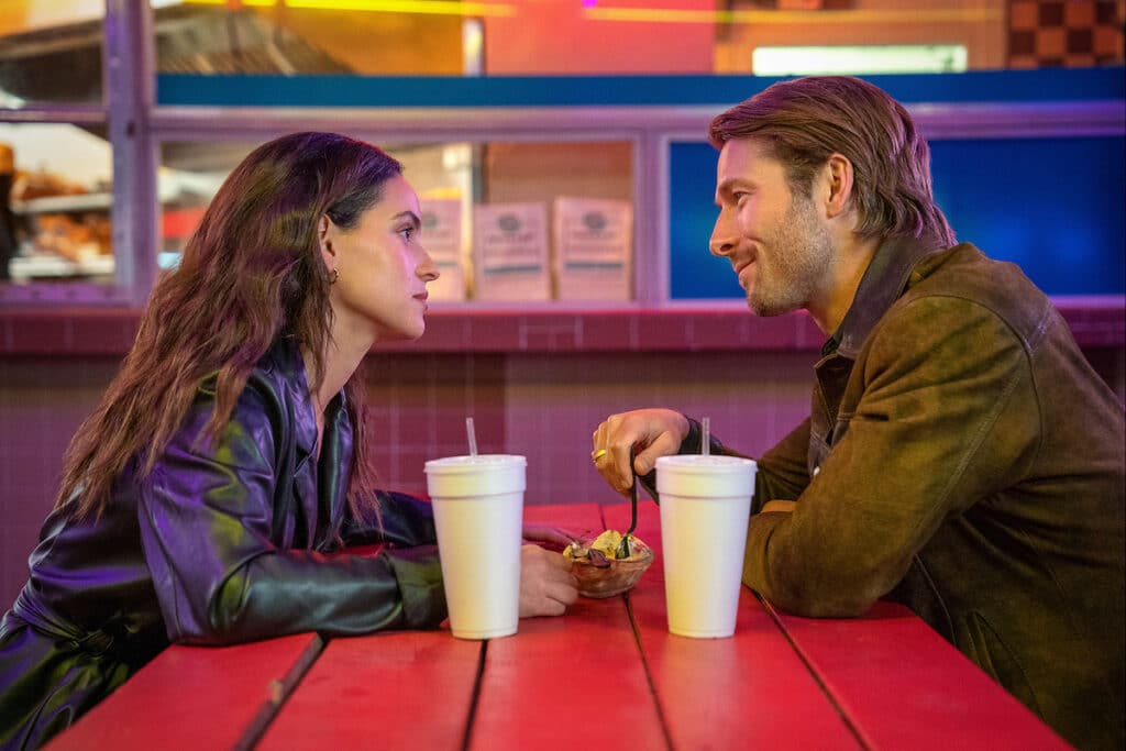 Adria Arjona and Glen Powell in a booth with big drinks with straws