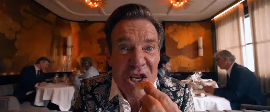 Dennis Quaid eating at a table, holding a shrimp