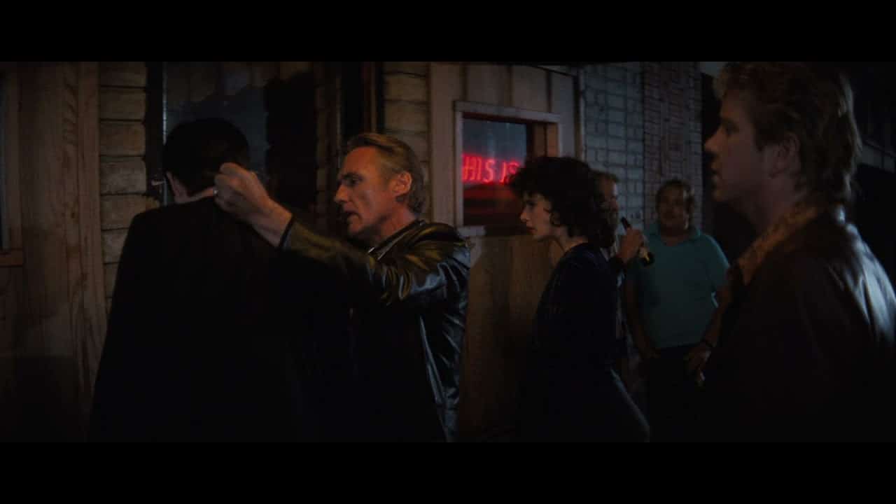 On a dark proch, Dennis Hopper grabs Kyle MacLachlan by the neck as friends watch