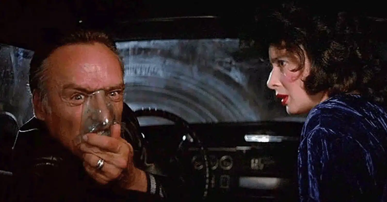 In the front seat of a car, Dennis Hopper with his gas mask on sitting next to Isabella Rossellini