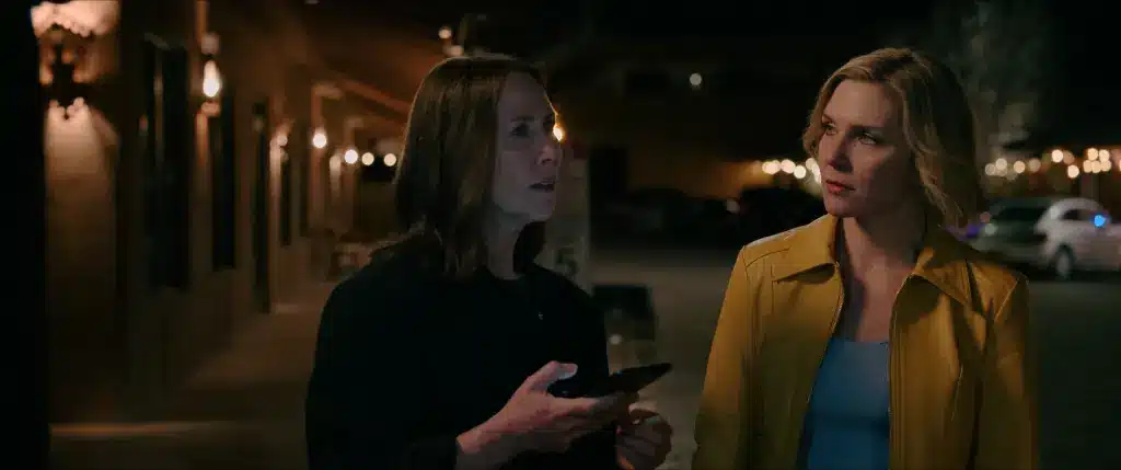 Helen holding a phone as she talks to Carol on the porch of a bar at night