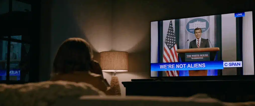 Carol sitting on her living room couch, watching the news on a big screen tv on the wall